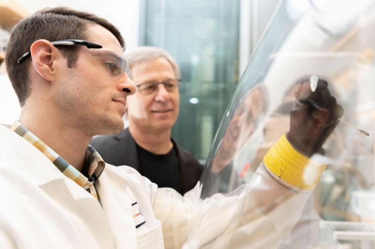 <p>Marcus Bray, front, observes a sample inside a sealed atmospheric tent that has no breathable oxygen. The tent simulates atmospheric gas mixtures during Earth's earliest eon and allows researchers to work with samples by slipping their hands into leak-proof gloves. In the background, co-principal investigator and NASA astrobiologist Loren Williams looks on in his lab at Georgia Tech. Credit: Allison Carter</p>