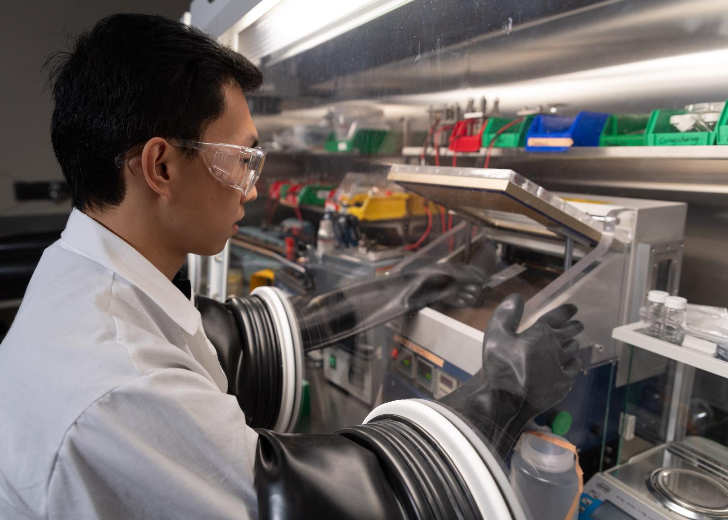 A scientist in a white lab coat uses protective equipment and rubber gloves to build a battery cell