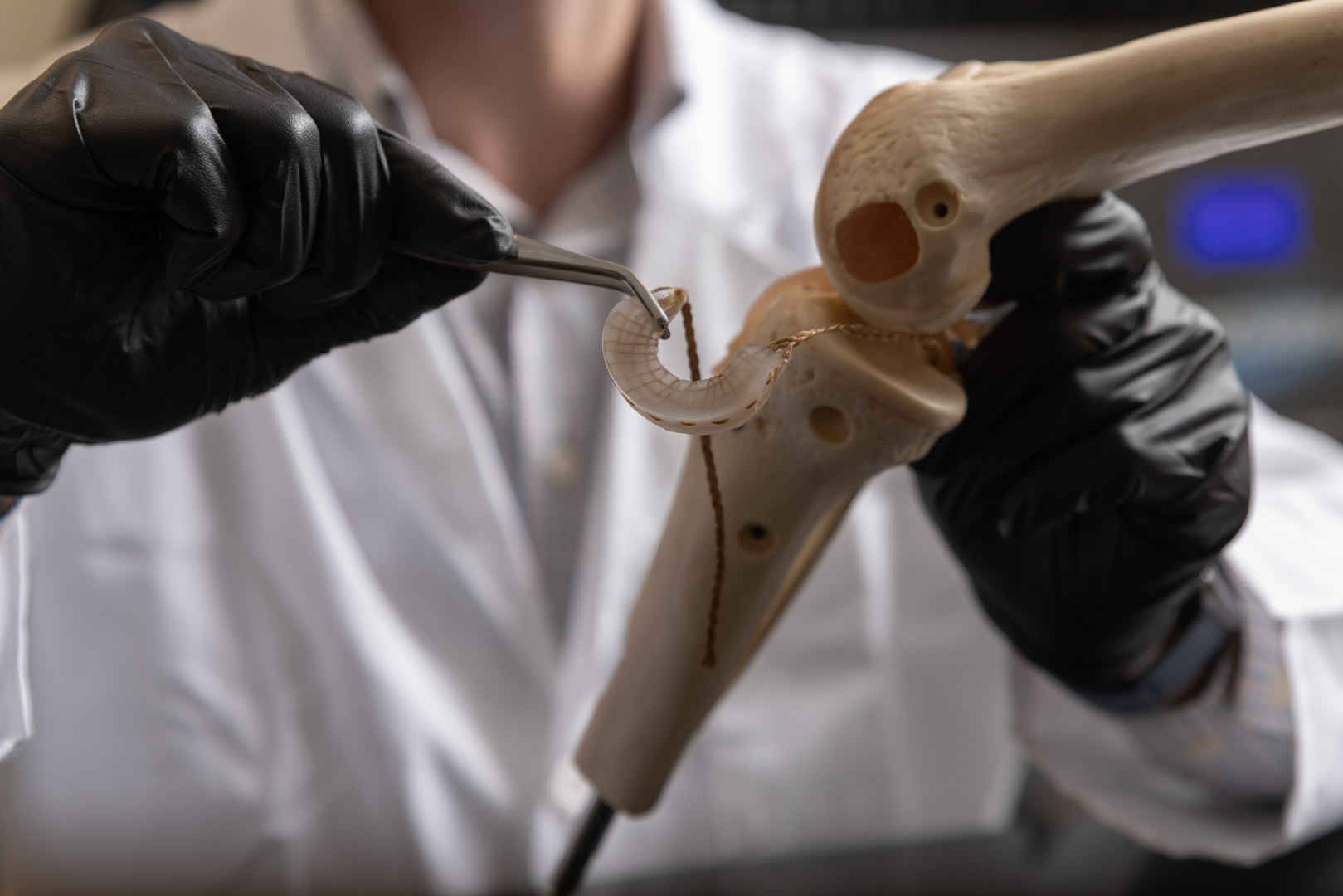 A figure with a white lab coat and black gloves interacts with a meniscus implant and a knee model. 