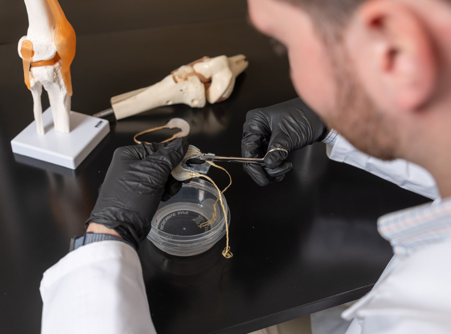 A figure in a white lab coat and black glove trims an implant. 