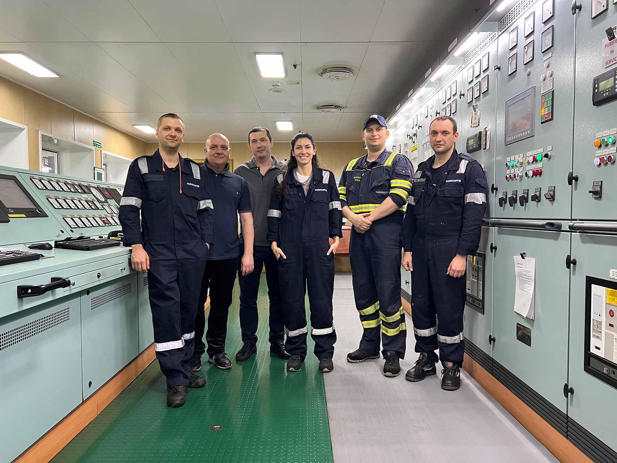 Six people in dark blue boiler suits standing in the control room of a ship