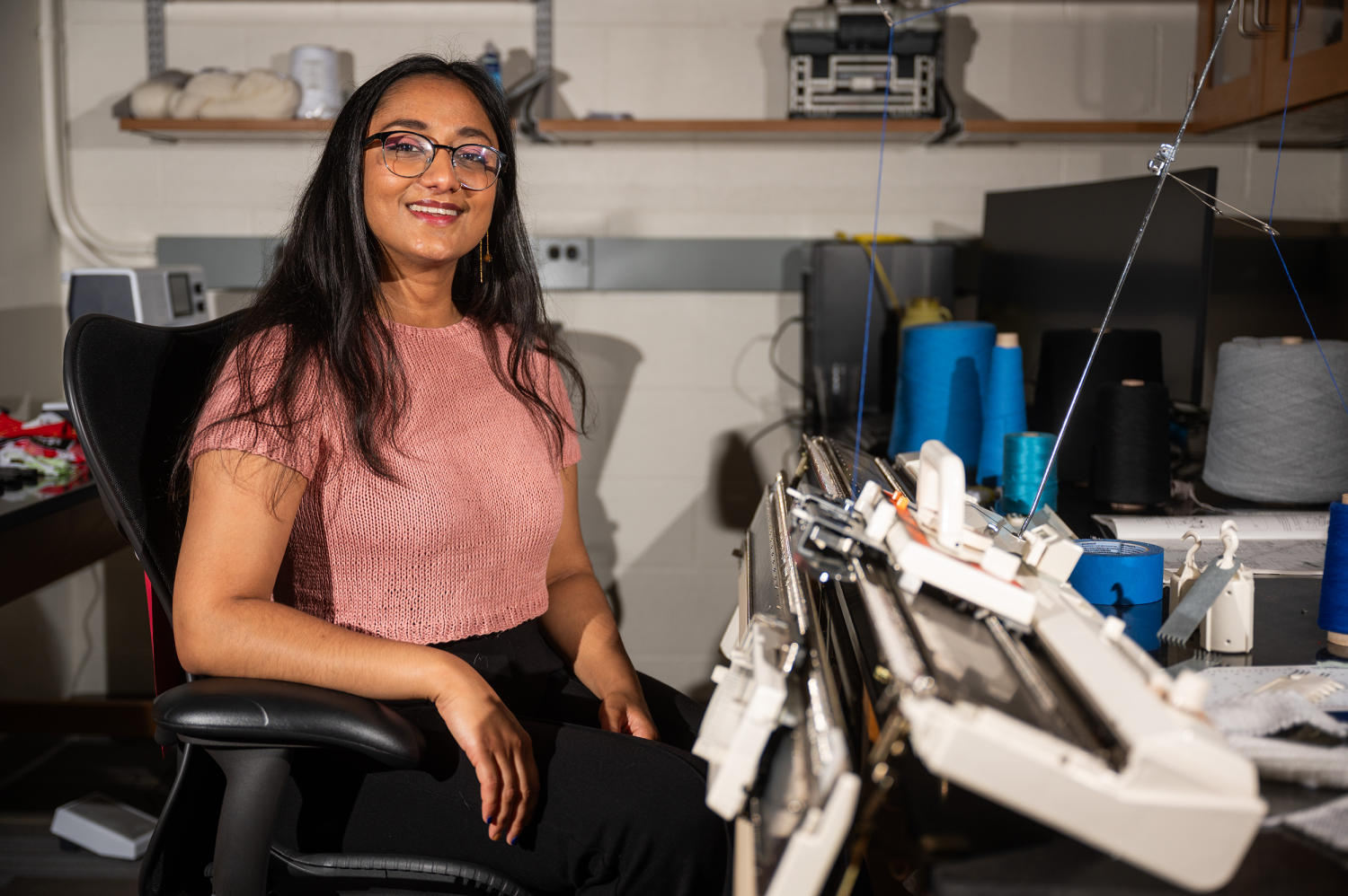 A woman wearing glasses and short sleeve pink sweater sit nexts to a commercial knitting machine.