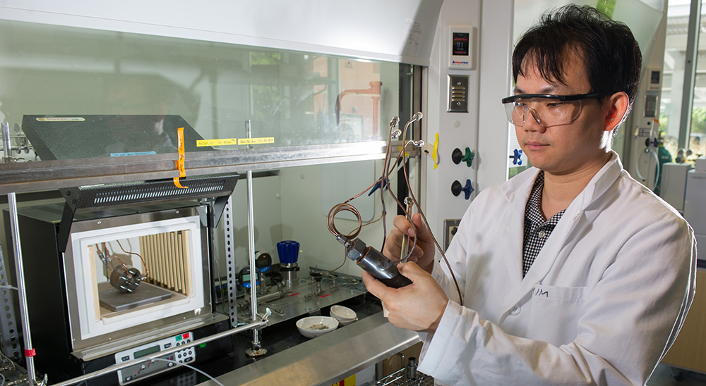 Man in a lab coat and safety glasses examines experimental hardware at a lab bench.