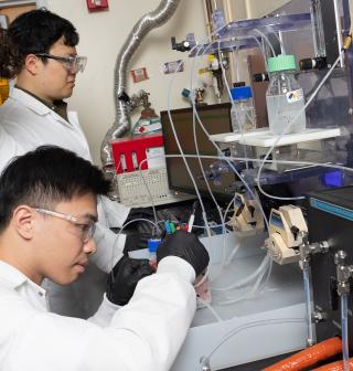 Three men in lab coats working at a bench on an experimental setup with tubes, vials, and pumps.