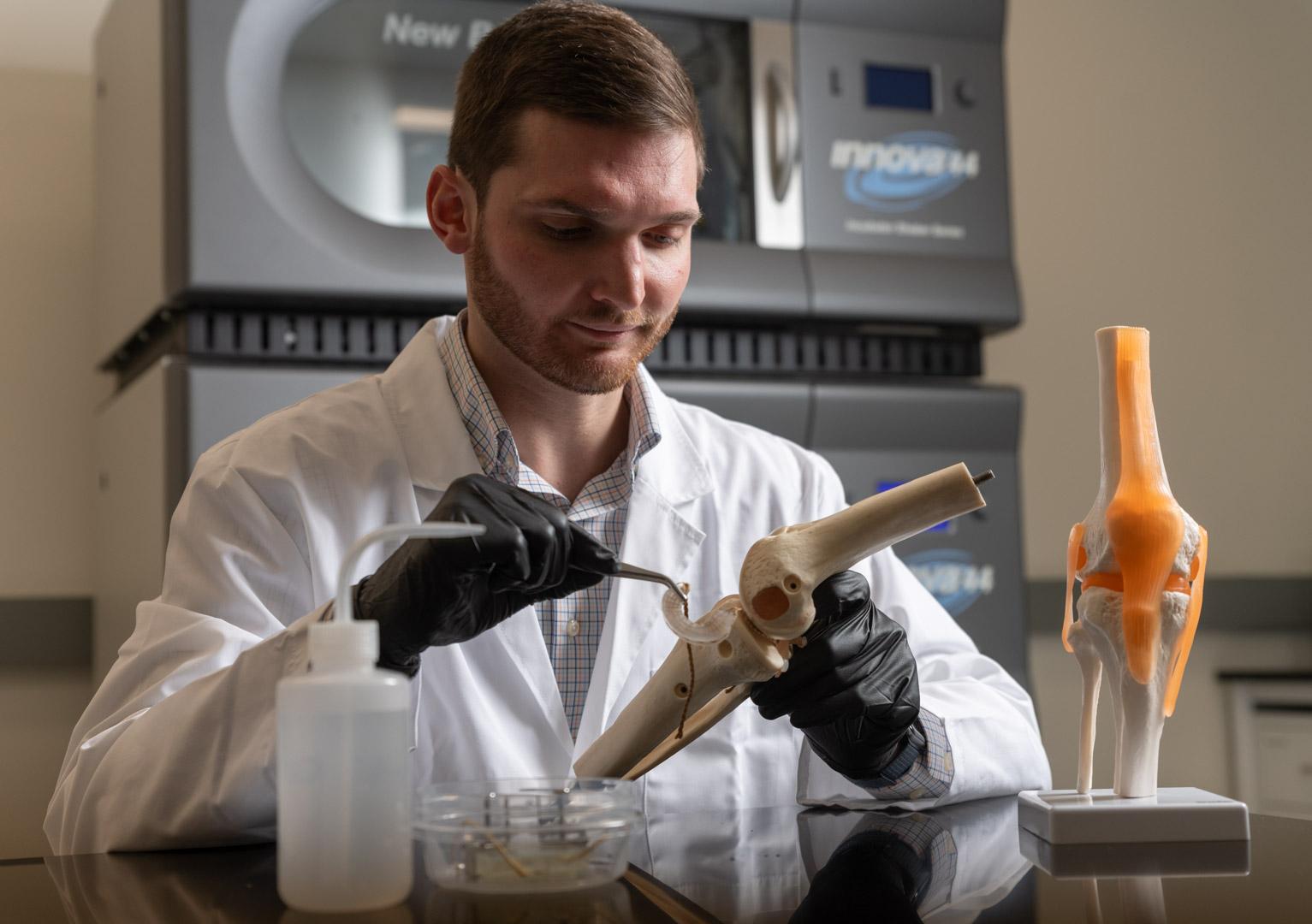 A man in a white lab coat and black gloves interacts with an implant and a knee model.